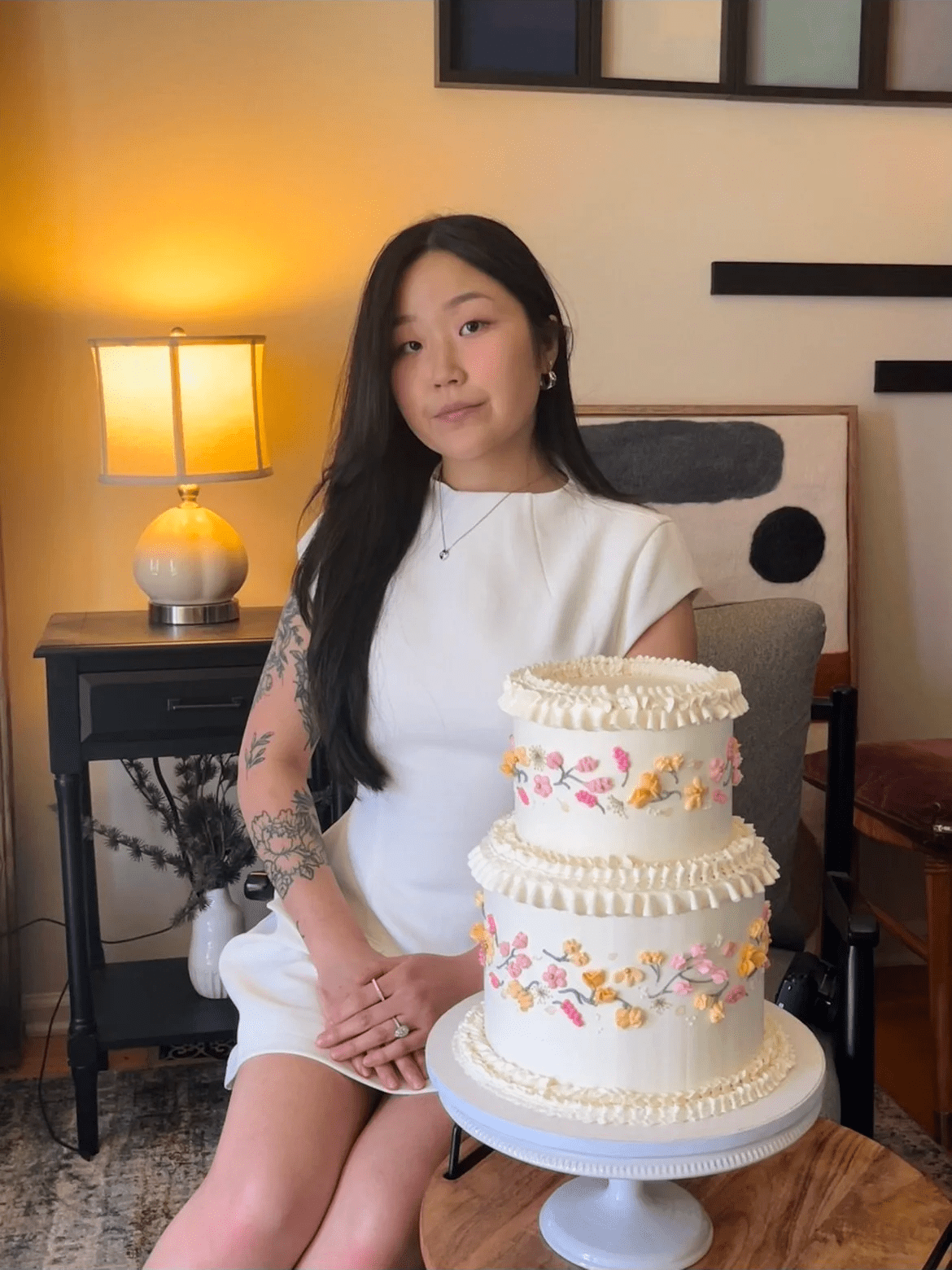 Young woman with long black hair sitting next to a decorated three-tier birthday cake with floral and butterfly designs on a cake stand, in a cozy indoor setting with a lamp, framed art, and a plant in the background.